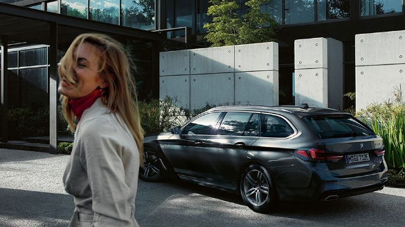 Frau lächelt, schwarzer BMW Kombi im Hintergrund, modernes Gebäude, sonniger Tag.