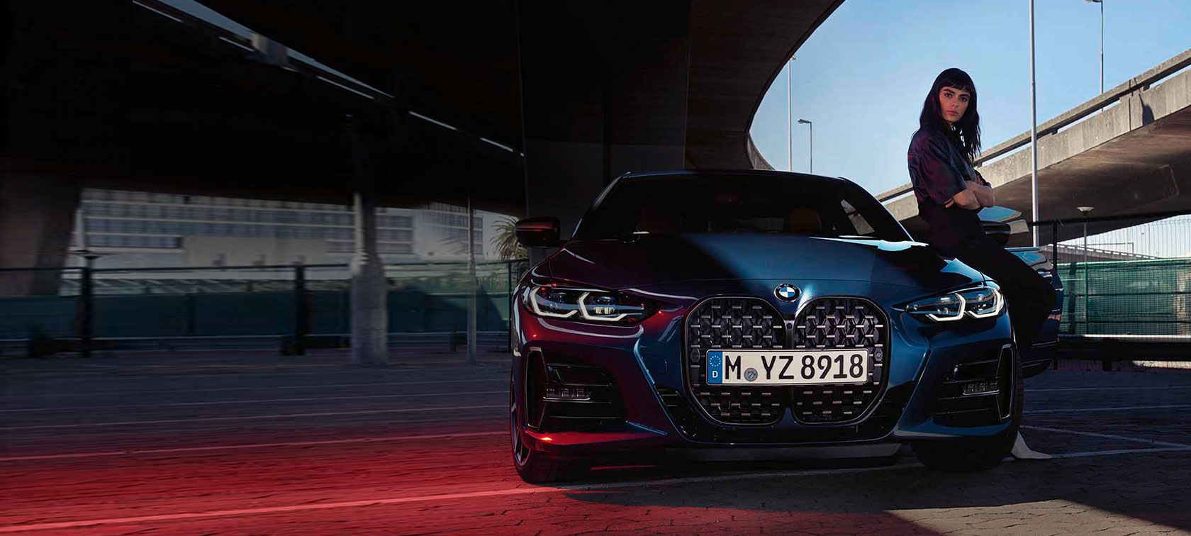 Blaues BMW 4er Coupé vor Brücke, Person sitzt auf Motorhaube, tagsüber.