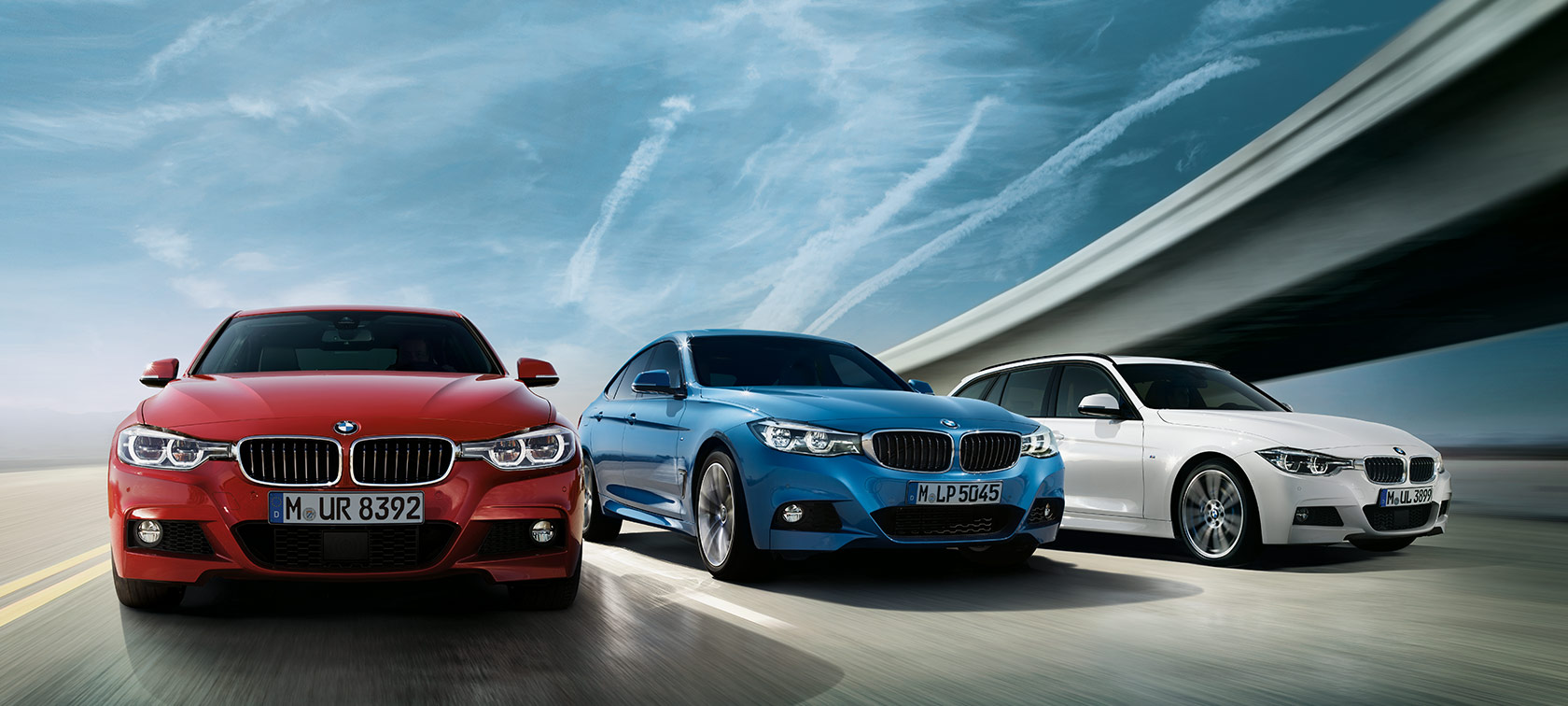 Drei BMWs in Rot, Blau und Weiß fahren auf einer Straße unter blauem Himmel.