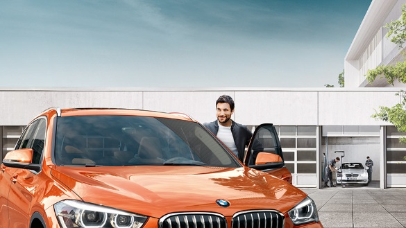 Ein Mann steht neben einem orangefarbenen BMW vor einer Werkstatt; im Hintergrund ein weiterer BMW.