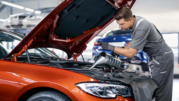 Ein Mechaniker füllt Öl in einen geöffneten Motorraum eines roten Autos in einer Werkstatt.