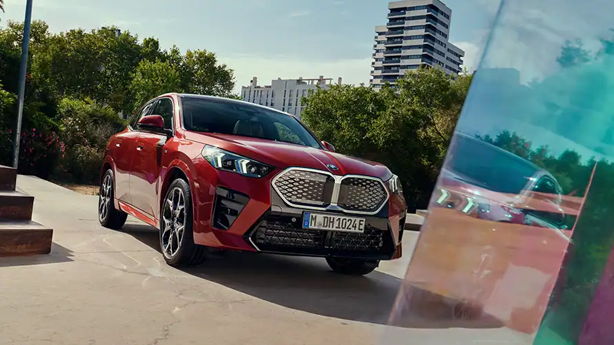 Roter BMW iX2 parkt draußen vor Bäumen und Gebäuden, Spiegelung im Vordergrund.