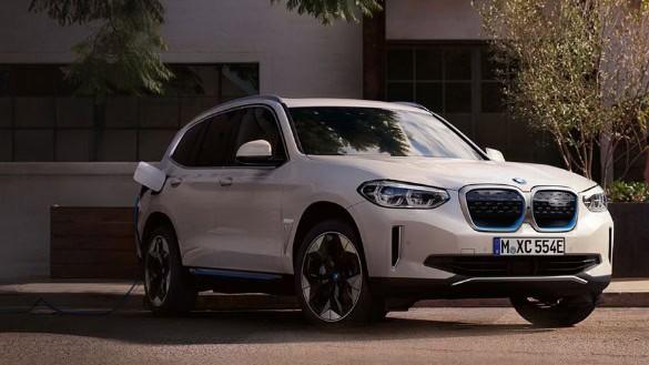 Weißer BMW iX3 SUV in städtischer Umgebung, seitlich von Bäumen beschattet.